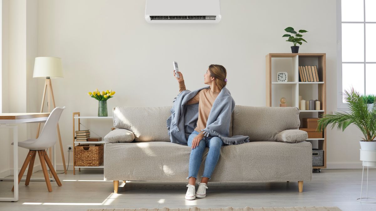 A lady with a blanket on her shoulders is sitting on a sofa and turning on the AC